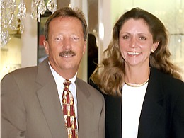 Two professionally dressed people smiling, standing together in a well-lit indoor setting.